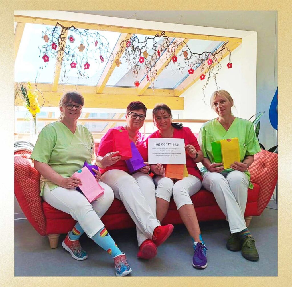 Vier Pflegekräfte in bunter Kleidung sitzen auf einem Sofa und halten eine Dankesurkunde zum Tag der Pflege in die Kamera
